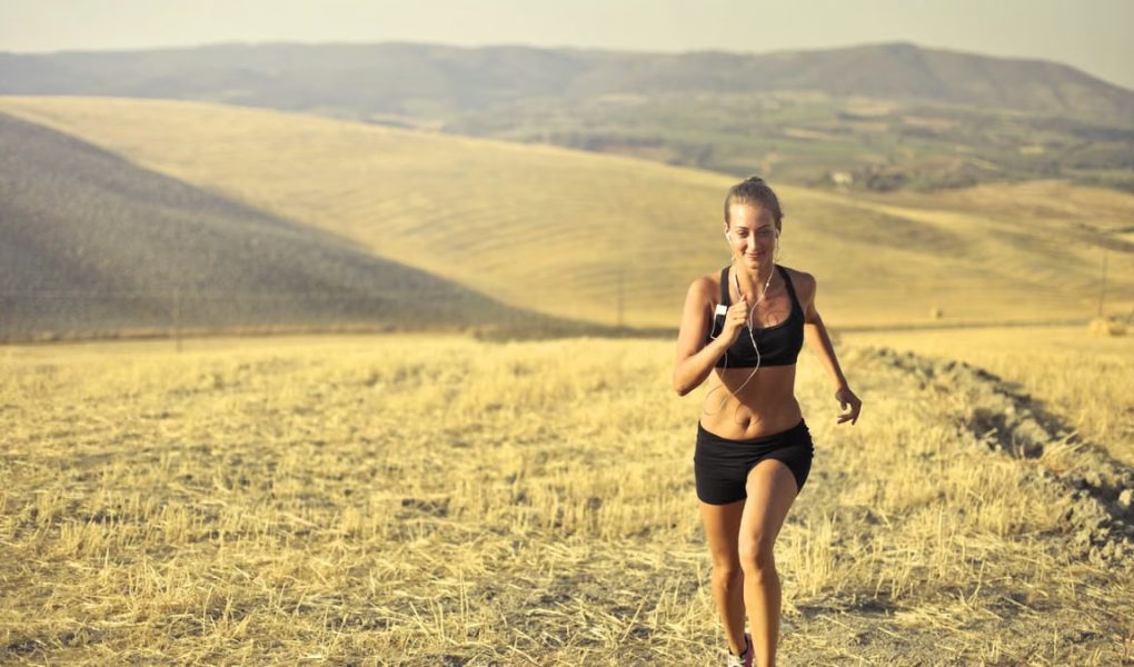 Powerful young female athlete in activewear running along hill on background of mountainous landscape and listening to music in earphones during cardio training