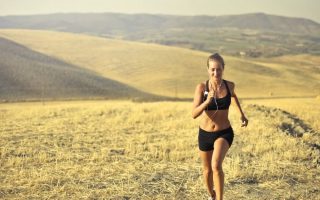 Powerful young female athlete in activewear running along hill on background of mountainous landscape and listening to music in earphones during cardio training