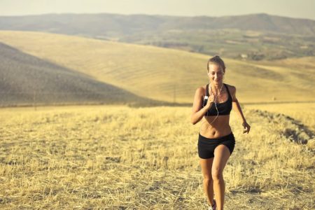Powerful young female athlete in activewear running along hill on background of mountainous landscape and listening to music in earphones during cardio training